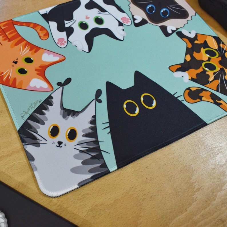 May include: A rectangular mouse mat with a light teal background and a playful design of cartoon cats. The cats are in various colours, including orange, black, grey, and calico, with large, expressive eyes. The mouse mat has a white stitched edge.