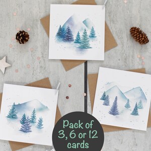May include: Three watercolour greeting cards with a winter mountain scene. The cards feature blue and white mountains with green trees. The text "Pack of 3, 6 or 12 cards" is printed on a black background.