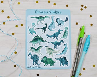 May include: A sheet of "Dinosaur Stickers" featuring illustrations of dinosaurs in blue and green hues. Two pens, one green and one blue, are placed beside the stickers. Gold confetti and blue and white string are scattered around the sheet.