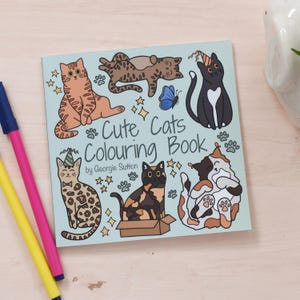 May include: A blue and white colouring book with the title "Cute Cats Colouring Book" by Georgie Sutton. The cover features illustrations of various cats in different poses, including a cat wearing a party hat, a cat sleeping, and a cat sitting in a box.