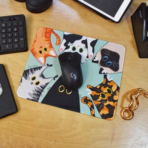 May include: A rectangular mouse mat with a light blue background and cartoon cat illustrations in various colours. A black wireless mouse sits on the mat. The cats include orange, black, calico, and Siamese cat designs. The mouse mat is on a wooden desk.