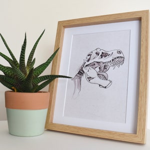 Puede incluir: Una impresi&oacute;n enmarcada con una ilustraci&oacute;n en blanco y negro de un cr&aacute;neo de Tyrannosaurus Rex. La obra de arte est&aacute; en un marco de madera clara. A la izquierda del marco hay una planta suculenta en maceta con hojas verdes y marrones.