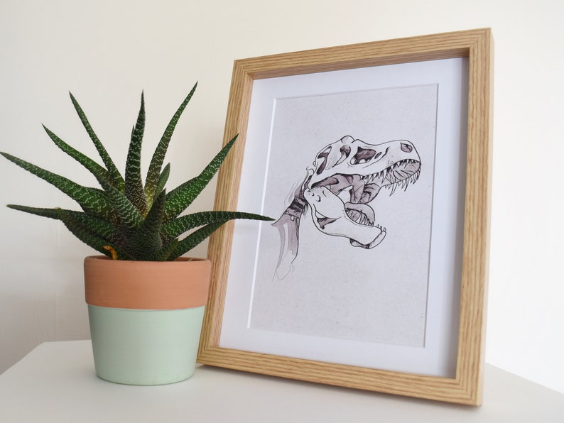 Puede incluir: Una impresi&oacute;n enmarcada con una ilustraci&oacute;n en blanco y negro de un cr&aacute;neo de Tyrannosaurus Rex. La obra de arte est&aacute; en un marco de madera clara. A la izquierda del marco hay una planta suculenta en maceta con hojas verdes y marrones.