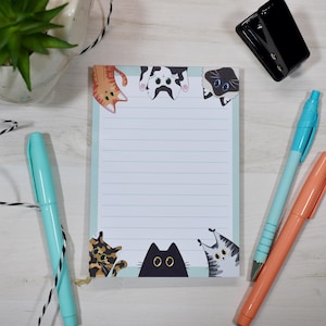 May include: A lined notepad with a light blue border and cat illustrations. The notepad features orange, black, and grey cats at the top and bottom. A black hole punch, a teal pen, and an orange pen are also visible.