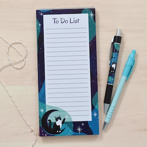 Peut inclure: Un bloc-notes de liste de choses à faire avec un motif de chat noir et blanc sur un fond de galaxie bleu et violet. Le bloc-notes a un fond blanc avec des lignes noires pour écrire.
