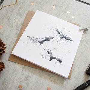 Peut inclure: Une carte blanche avec une illustration en noir et blanc de trois chauves-souris volant sur un fond de style aquarelle. La carte est sur une surface brune.