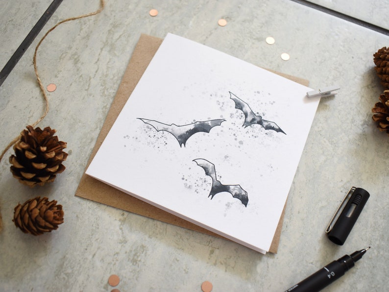 Peut inclure: Une carte blanche avec une illustration en noir et blanc de trois chauves-souris volant sur un fond de style aquarelle. La carte est sur une surface brune.