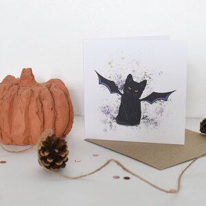 Peut inclure: Une carte de v&oelig;ux repr&eacute;sentant un chat noir avec des ailes de chauve-souris violettes sur un fond blanc avec des &eacute;claboussures d'aquarelle violettes et vertes. La carte est vierge &agrave; l'int&eacute;rieur et est livr&eacute;e avec une enveloppe brune.