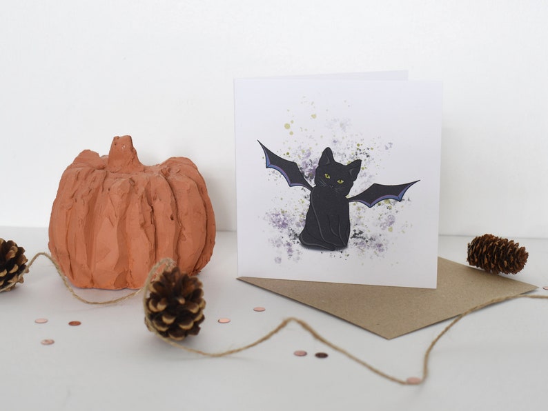 Peut inclure: Une carte de v&oelig;ux repr&eacute;sentant un chat noir avec des ailes de chauve-souris violettes sur un fond blanc avec des &eacute;claboussures d'aquarelle violettes et vertes. La carte est vierge &agrave; l'int&eacute;rieur et est livr&eacute;e avec une enveloppe brune.