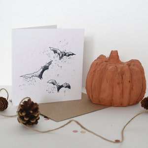 Peut inclure: Une carte blanche avec une illustration en noir et blanc de chauves-souris volant dans un ciel nuageux. La carte est pos&eacute;e sur une enveloppe brune avec une petite citrouille orange et des pommes de pin sur une surface blanche.