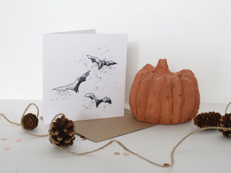 Peut inclure: Une carte blanche avec une illustration en noir et blanc de chauves-souris volant dans un ciel nuageux. La carte est pos&eacute;e sur une enveloppe brune avec une petite citrouille orange et des pommes de pin sur une surface blanche.