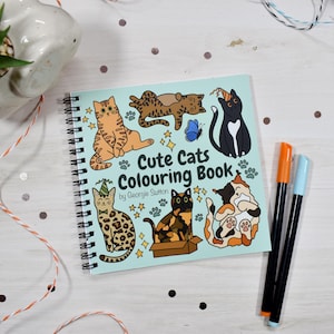 May include: A spiral-bound coloring book titled "Cute Cats Colouring Book" by Georgie Sutton. The cover features cartoon cats in various poses, with stars and a blue butterfly. Two markers, orange and blue, are next to the book.