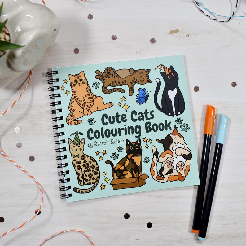 May include: A spiral-bound coloring book titled "Cute Cats Colouring Book" by Georgie Sutton. The cover features cartoon cats in various poses, with stars and a blue butterfly. Two markers, orange and blue, are next to the book.