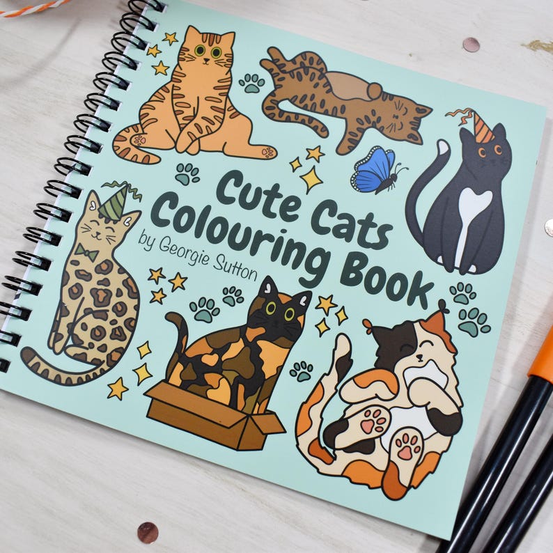 May include: A spiral-bound coloring book titled "Cute Cats Colouring Book" by Georgie Sutton. The cover features cartoon illustrations of various cats, paw prints, stars, and a blue butterfly. The cats are in different poses and colors.