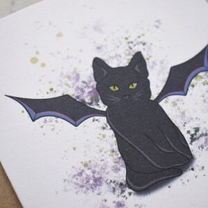 Peut inclure: Un chat noir aux yeux jaunes et des ailes de chauve-souris assis sur un fond blanc avec des &eacute;claboussures d'aquarelle violettes et vertes.