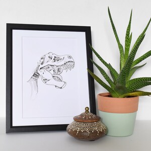 Puede incluir: Ilustraci&oacute;n en blanco y negro enmarcada de un cr&aacute;neo de Tyrannosaurus Rex. Un peque&ntilde;o tarro de cer&aacute;mica marr&oacute;n decorativo con tapa se encuentra frente al marco. A la derecha hay una planta suculenta en maceta con hojas verdes y amarillas.
