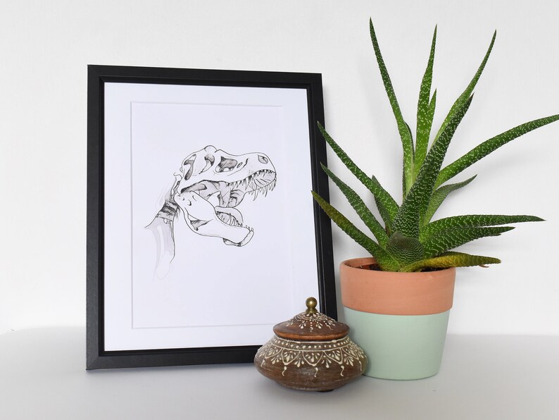 Puede incluir: Ilustraci&oacute;n en blanco y negro enmarcada de un cr&aacute;neo de Tyrannosaurus Rex. Un peque&ntilde;o tarro de cer&aacute;mica marr&oacute;n decorativo con tapa se encuentra frente al marco. A la derecha hay una planta suculenta en maceta con hojas verdes y amarillas.