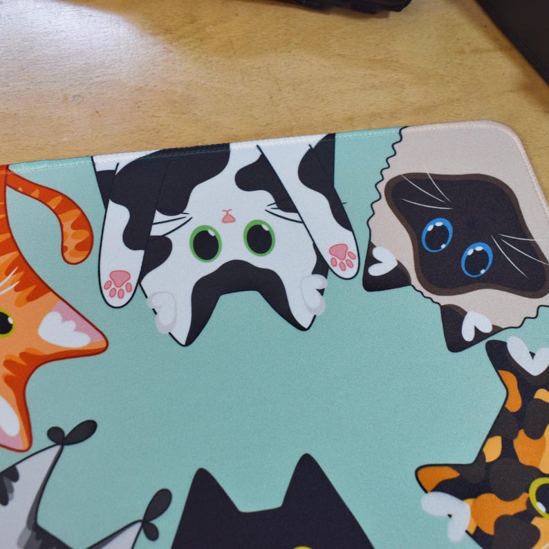 May include: A rectangular mouse mat with a light teal background features cartoon cat faces in various colours. The cats include orange, black, white, and Siamese. The mat is designed for computer use.