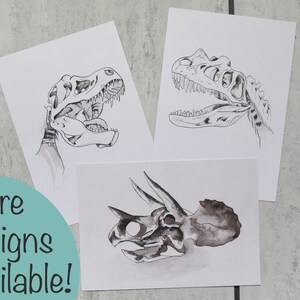 Puede incluir: Tres impresiones art&iacute;sticas de calaveras de dinosaurios. Las impresiones presentan ilustraciones detalladas en blanco y negro de una calavera de T-Rex y una calavera de Triceratops. El texto "More designs available!" est&aacute; en un c&iacute;rculo turquesa.
