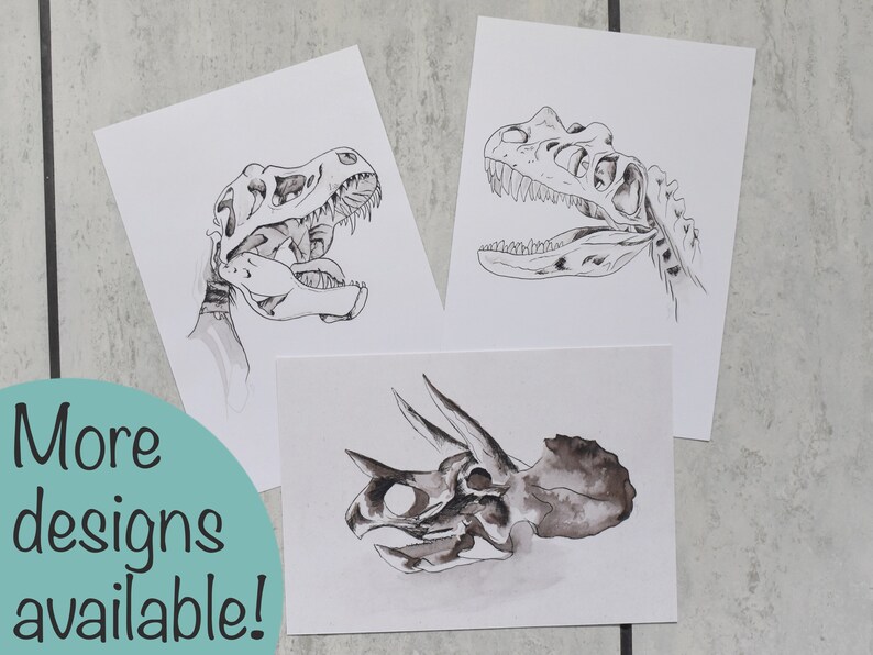 Puede incluir: Tres impresiones art&iacute;sticas de calaveras de dinosaurios. Las impresiones presentan ilustraciones detalladas en blanco y negro de una calavera de T-Rex y una calavera de Triceratops. El texto "More designs available!" est&aacute; en un c&iacute;rculo turquesa.