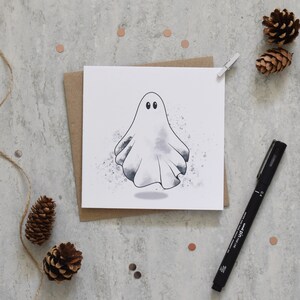 Peut inclure: Un fant&ocirc;me blanc avec deux yeux noirs est dessin&eacute; sur une carte blanche. La carte est sur une enveloppe brune. L'arri&egrave;re-plan est une surface grise avec des pommes de pin brunes et de petits points de couleur cuivre.
