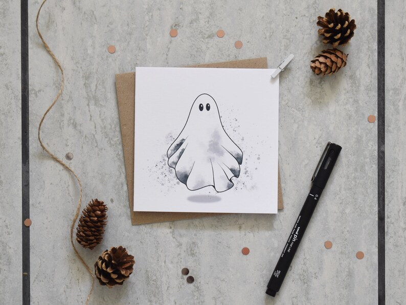 Peut inclure: Un fant&ocirc;me blanc avec deux yeux noirs est dessin&eacute; sur une carte blanche. La carte est sur une enveloppe brune. L'arri&egrave;re-plan est une surface grise avec des pommes de pin brunes et de petits points de couleur cuivre.
