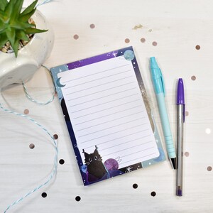 May include: A lined notepad with a space-themed design featuring a black cat, planets, and stars. A light blue pen and a purple pen are next to the notepad. A small succulent plant in a white pot is in the background.
