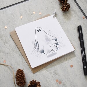 Peut inclure: Un fant&ocirc;me blanc avec des yeux noirs est dessin&eacute; sur une carte blanche avec un fond aquarelle gris. La carte est sur une enveloppe brune.