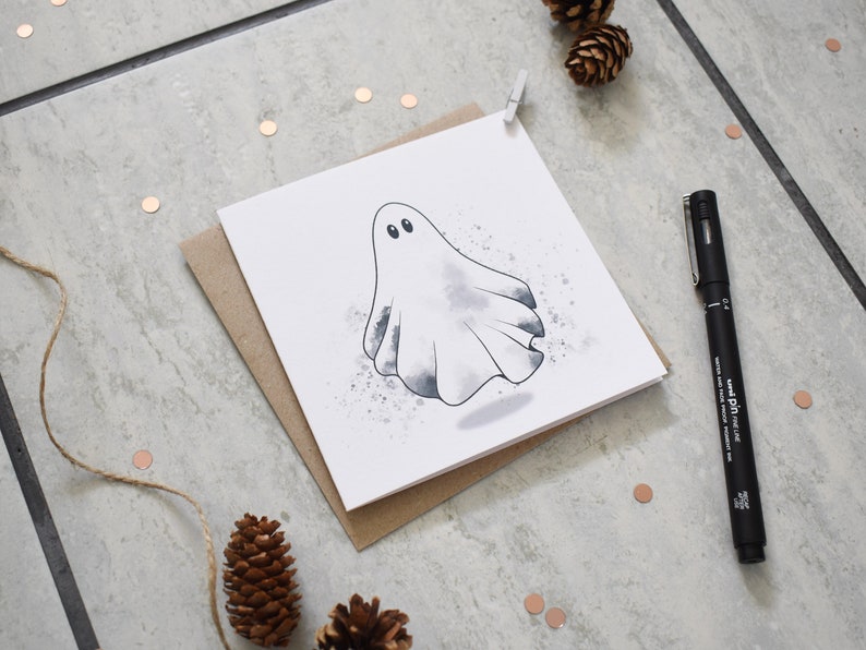 Peut inclure: Un fant&ocirc;me blanc avec des yeux noirs est dessin&eacute; sur une carte blanche avec un fond aquarelle gris. La carte est sur une enveloppe brune.