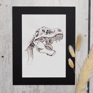 Puede incluir: Un dibujo a tinta en blanco y negro de un cr&aacute;neo de Tyrannosaurus Rex, enmarcado en un passe-partout negro. La obra de arte presenta sombreado y l&iacute;neas detalladas, mostrando los dientes y la estructura &oacute;sea del dinosaurio. La obra se exhibe sobre una superficie gris claro.