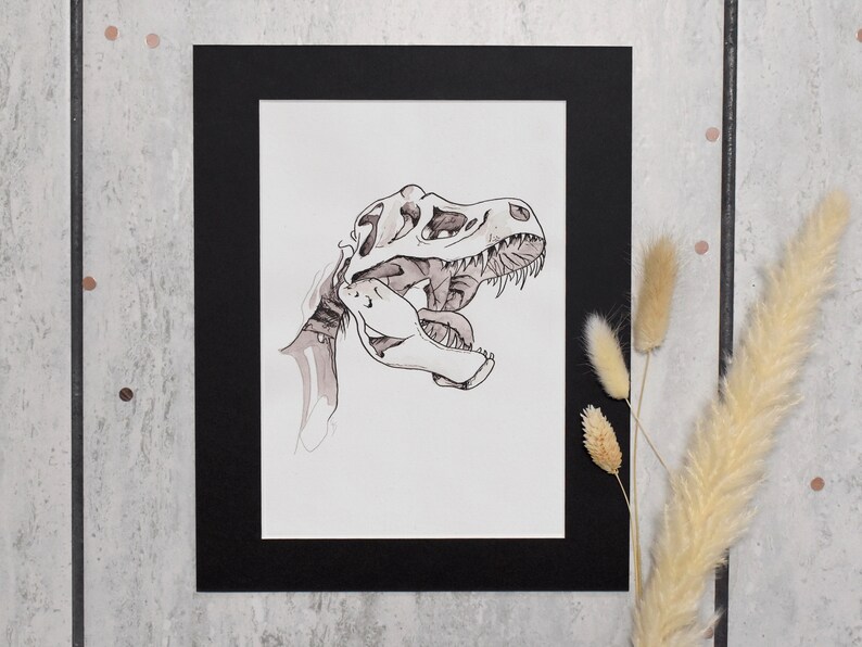 Puede incluir: Un dibujo a tinta en blanco y negro de un cr&aacute;neo de Tyrannosaurus Rex, enmarcado en un passe-partout negro. La obra de arte presenta sombreado y l&iacute;neas detalladas, mostrando los dientes y la estructura &oacute;sea del dinosaurio. La obra se exhibe sobre una superficie gris claro.
