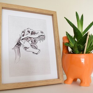 Puede incluir: Ilustraci&oacute;n en blanco y negro enmarcada de un cr&aacute;neo de Tyrannosaurus Rex. La obra de arte est&aacute; en un marco de madera clara. A la derecha hay una maceta naranja de cer&aacute;mica con forma de dinosaurio con una planta suculenta verde.