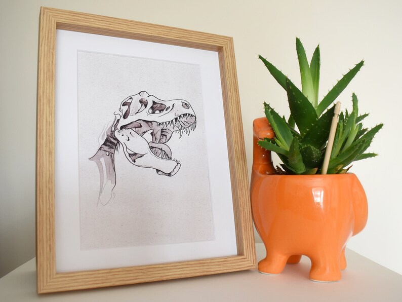 Puede incluir: Ilustraci&oacute;n en blanco y negro enmarcada de un cr&aacute;neo de Tyrannosaurus Rex. La obra de arte est&aacute; en un marco de madera clara. A la derecha hay una maceta naranja de cer&aacute;mica con forma de dinosaurio con una planta suculenta verde.