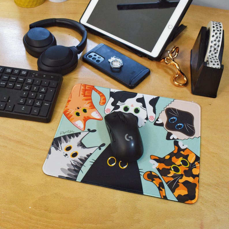 May include: A rectangular mouse mat featuring cartoon cats in various colours on a light blue background. A black computer mouse is on the mat, alongside other office items.