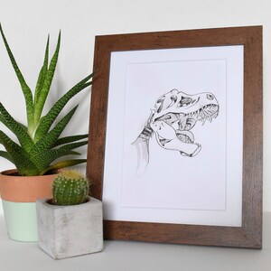 Puede incluir: Ilustraci&oacute;n en blanco y negro enmarcada de un cr&aacute;neo de Tyrannosaurus Rex. La obra de arte est&aacute; en un marco de madera marr&oacute;n oscuro, con un passe-partout blanco. Una suculenta y un cactus en maceta est&aacute;n en primer plano.