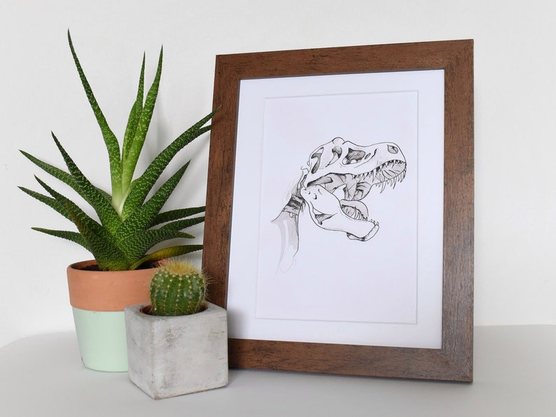 Puede incluir: Ilustraci&oacute;n en blanco y negro enmarcada de un cr&aacute;neo de Tyrannosaurus Rex. La obra de arte est&aacute; en un marco de madera marr&oacute;n oscuro, con un passe-partout blanco. Una suculenta y un cactus en maceta est&aacute;n en primer plano.