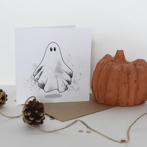 Peut inclure: Une carte blanche avec un fant&ocirc;me blanc aux yeux noirs et un fond aquarelle gris. La carte est pos&eacute;e sur une enveloppe brune avec une petite citrouille et des pommes de pin sur une surface blanche.