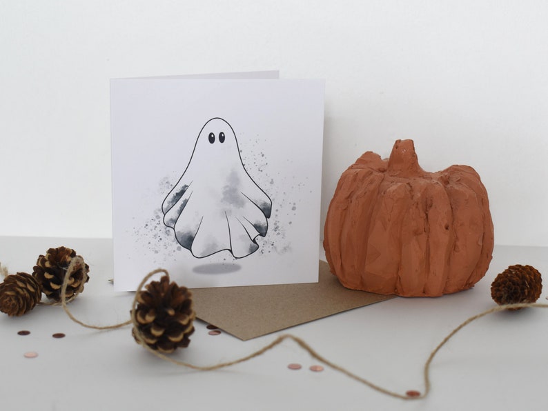 Peut inclure: Une carte blanche avec un fant&ocirc;me blanc aux yeux noirs et un fond aquarelle gris. La carte est pos&eacute;e sur une enveloppe brune avec une petite citrouille et des pommes de pin sur une surface blanche.