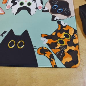 May include: A rectangular mouse mat with a light blue background and cartoon cat designs. The cats are black, tortoiseshell, and Siamese, with yellow and blue eyes. A black computer mouse is visible in the top right corner.