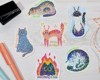 May include: A selection of cat-themed stickers with a shimmering, glittery finish. The stickers showcase diverse cat illustrations with text including "don't stress," "always on edge," and "everything is fine."