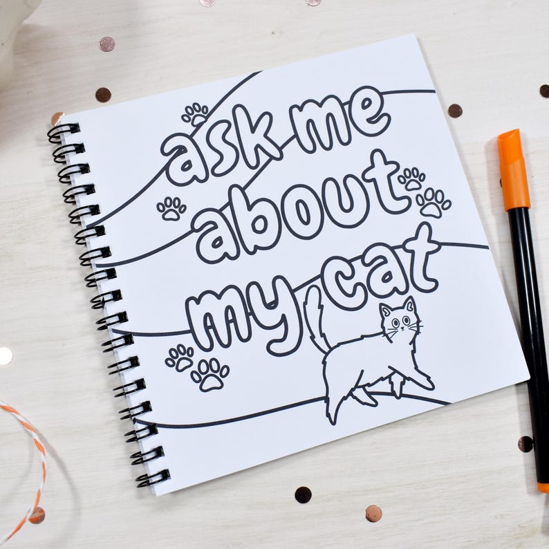 May include: A spiral-bound notebook with the text "ask me about my cat" and a cat illustration. The notebook is white with black text and a black spiral binding. An orange and black marker is next to the notebook.