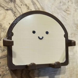 May include: A brown and beige phone stand shaped like a smiling piece of bread. The stand has two small arms on either side.