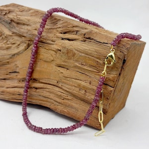 May include: A necklace composed of small, faceted, deep pinkish-red gemstone beads. The necklace features a gold-toned metal lobster claw clasp and an adjustable extender chain with elongated oval links, providing a secure and flexible closure.