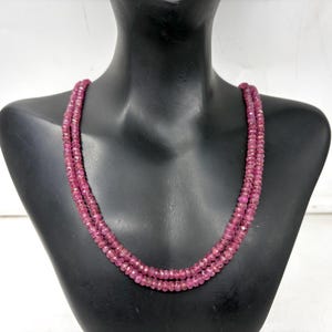 Puede incluir: Un collar de doble hebra compuesto por pequeñas cuentas facetadas de color rubí. El collar se muestra sobre un busto de maniquí negro, resaltando el color y el diseño de la joya. Las cuentas tienen un ligero brillo.