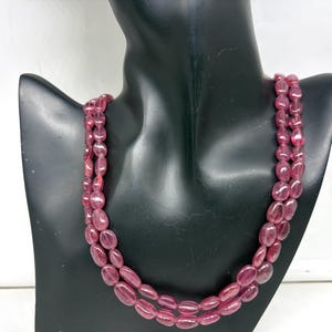 Può includere: Una collana a tre fili di pietre preziose rosa lucide di forma ovale. La collana è esposta su un busto di manichino nero. Le pietre preziose hanno una qualità traslucida e la collana è drappeggiata a cascata.