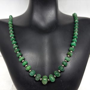 Può includere: Una collana di pietre preziose verdi vibranti e piccole perline color argento. La collana è esposta su un manichino nero, che mette in risalto il design e il colore della collana. Le pietre preziose hanno una forma arrotondata e sono infilate per formare una collana.