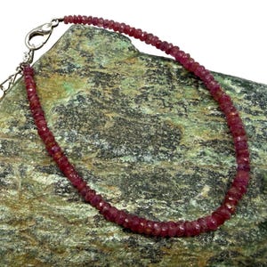 May include: A delicate ruby gemstone bracelet with a silver clasp. The bracelet is composed of small, faceted red gemstones. The jewelry is displayed on a textured, green and brown stone surface against a white background.