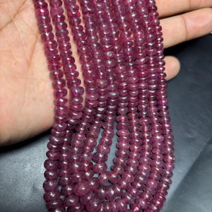 May include: A strand of pink ruby beads, approximately 4mm in size, with a smooth, polished finish. The beads are strung on a clear thread and are displayed on a hand.