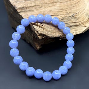 May include: A light blue beaded bracelet. The bracelet is made of round, polished beads. The beads are a soft, pastel blue color. The bracelet is displayed against a dark background with a piece of wood.