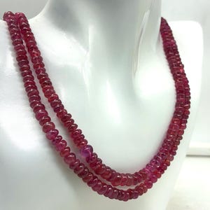 May include: A double strand necklace composed of small, round, deep red beads. The beads are strung together, creating a layered effect. The necklace is displayed on a white mannequin bust, highlighting the color and design of the jewelry.